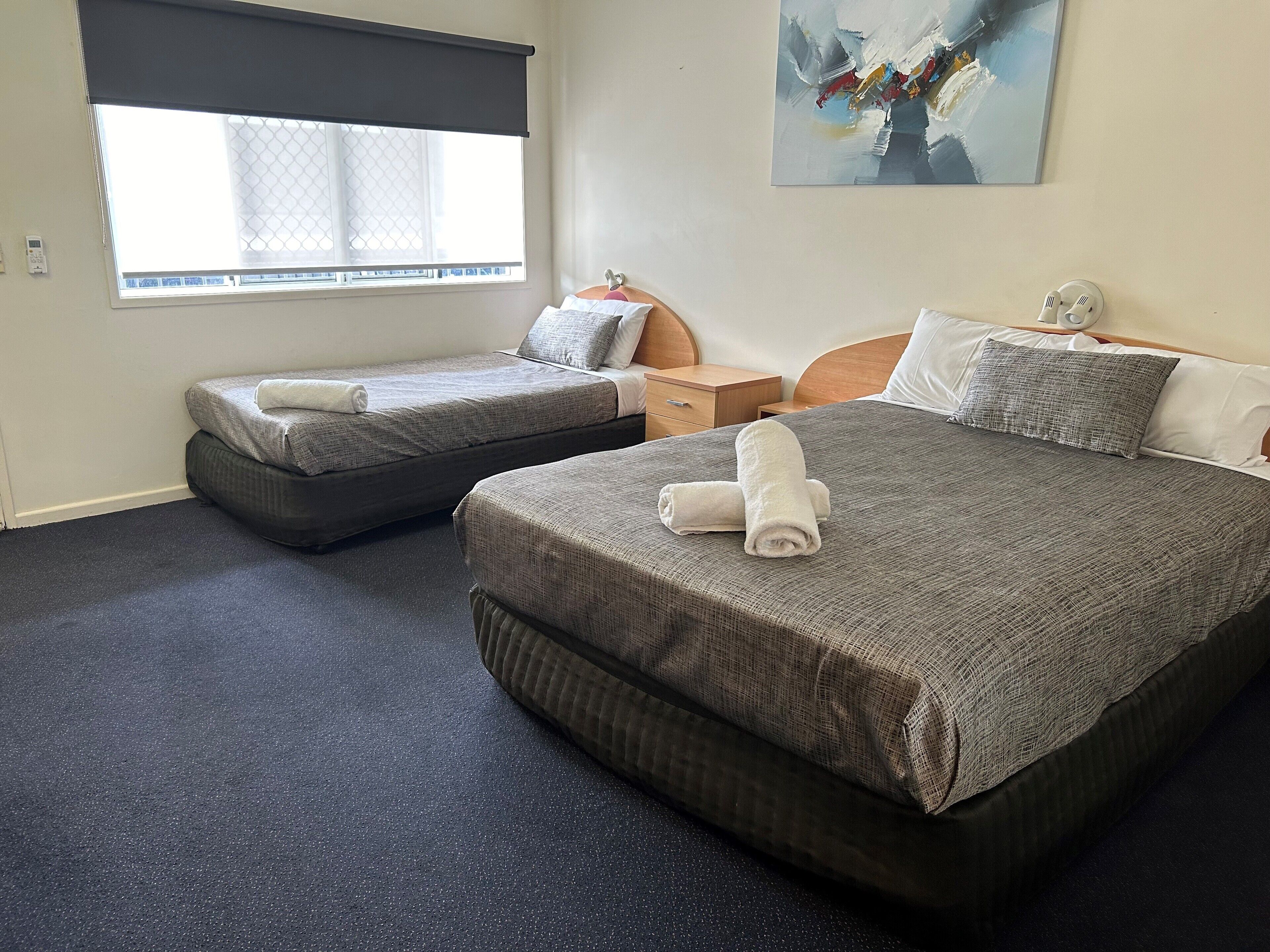Una habitación de hotel con dos camas, una ventana con cortina opaca, una mesita de noche y un cuadro en la pared.