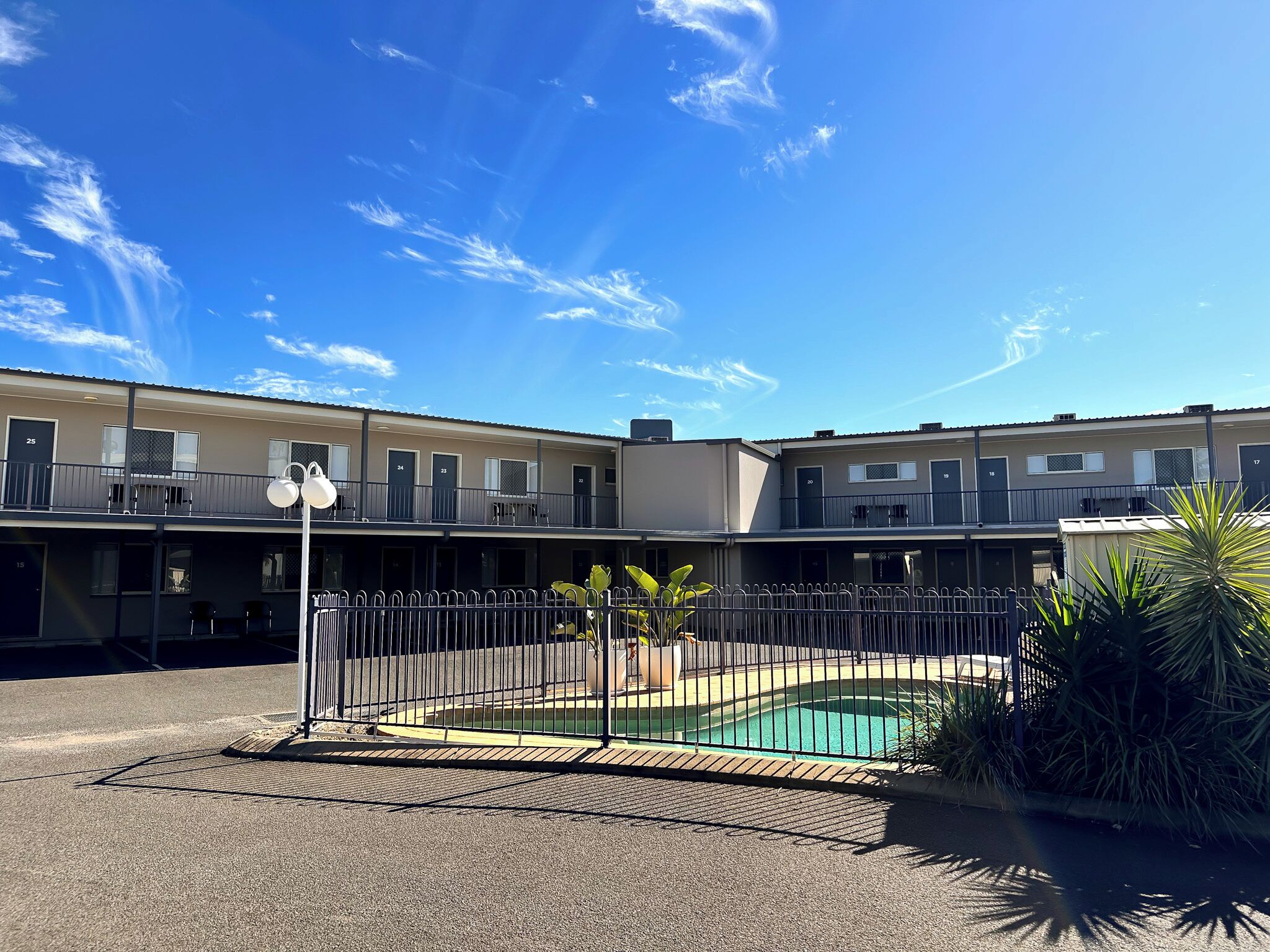 Un motel con zona de piscina, que incluye una piscina cercada, un pequeño jardín con plantas y un cielo azul despejado.