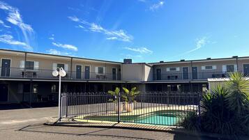 Un motel con zona de piscina, que incluye una piscina cercada, un pequeño jardín con plantas y un cielo azul despejado.