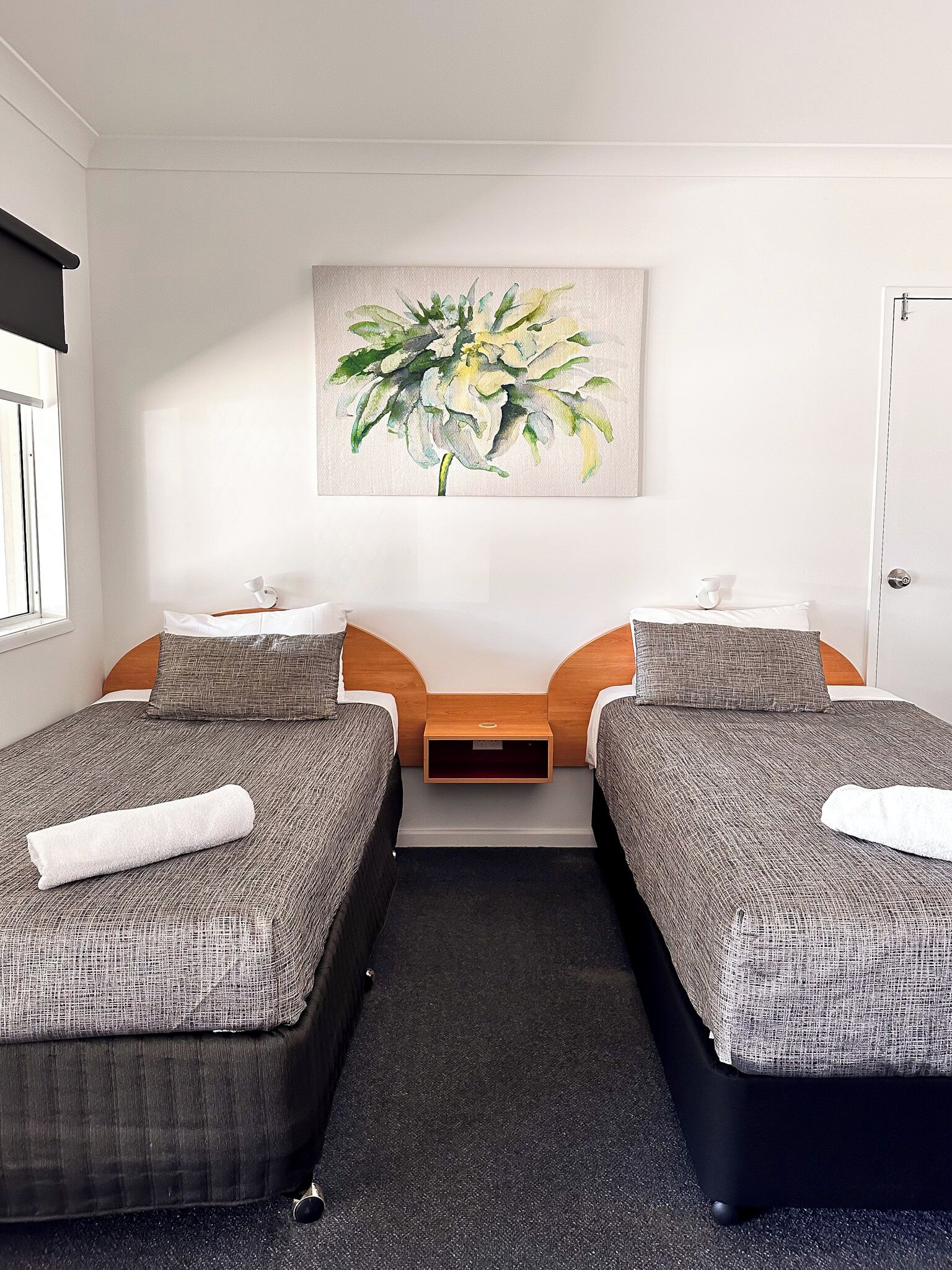 A hotel room with two single beds, a small bedside table, a painting on the wall, and a window with blinds.