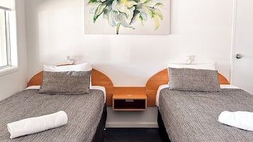 A hotel room with two single beds, a small bedside table, a painting on the wall, and a window with blinds.
