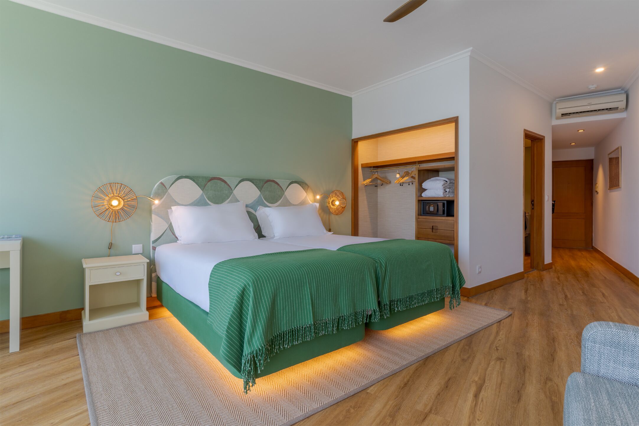 Une chambre à coucher avec un lit, des tables de chevet, une commode, une couverture verte et une étagère murale où sont rangés des vêtements.