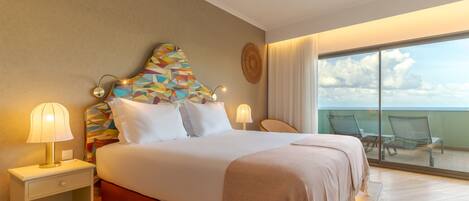 A hotel room with a large bed, two bedside tables with lamps, a colorful headboard, and a balcony with chairs.