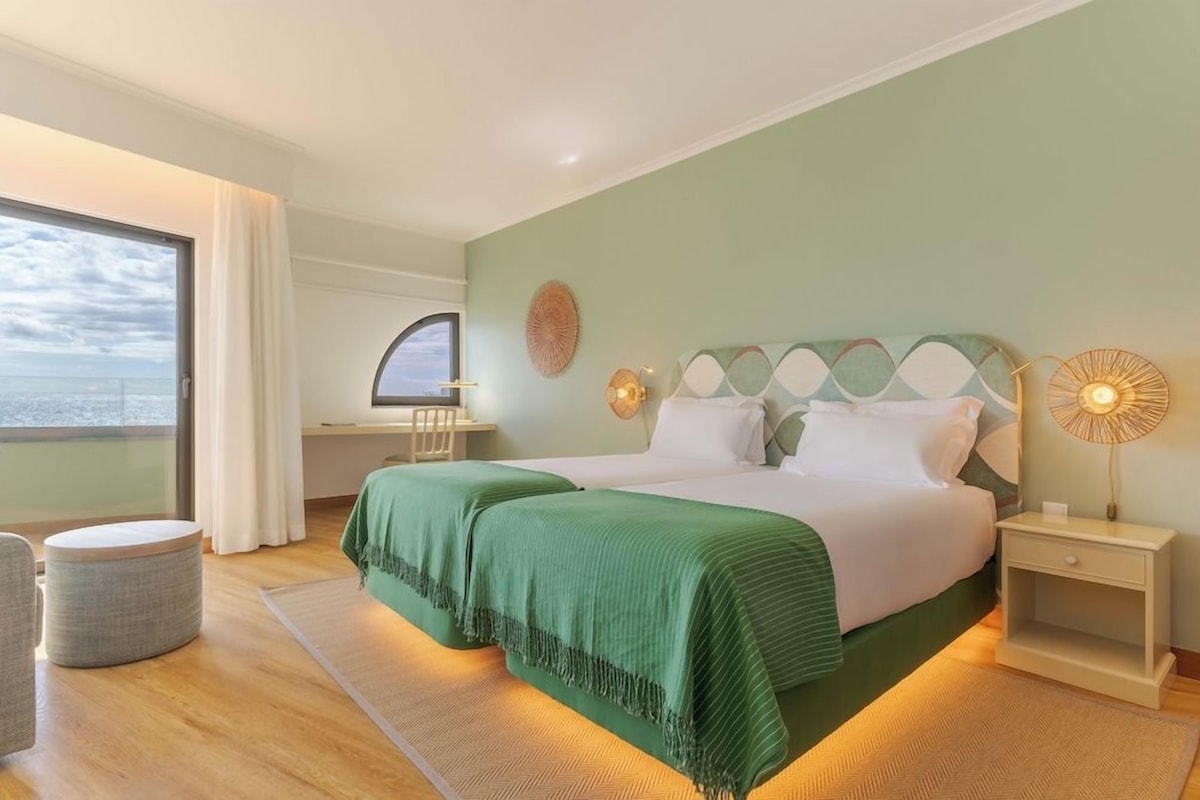 A hotel room with a bed, bedside tables, a chair, a window with a view, and a decorative wall piece.