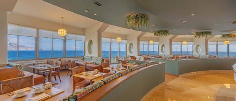 Um restaurante moderno com área de estar curvada, grandes janelas com vista para o oceano e plantas penduradas.