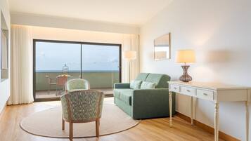 A room with a green sofa, a white desk, and a chair. There is a large window with a view of the sea.