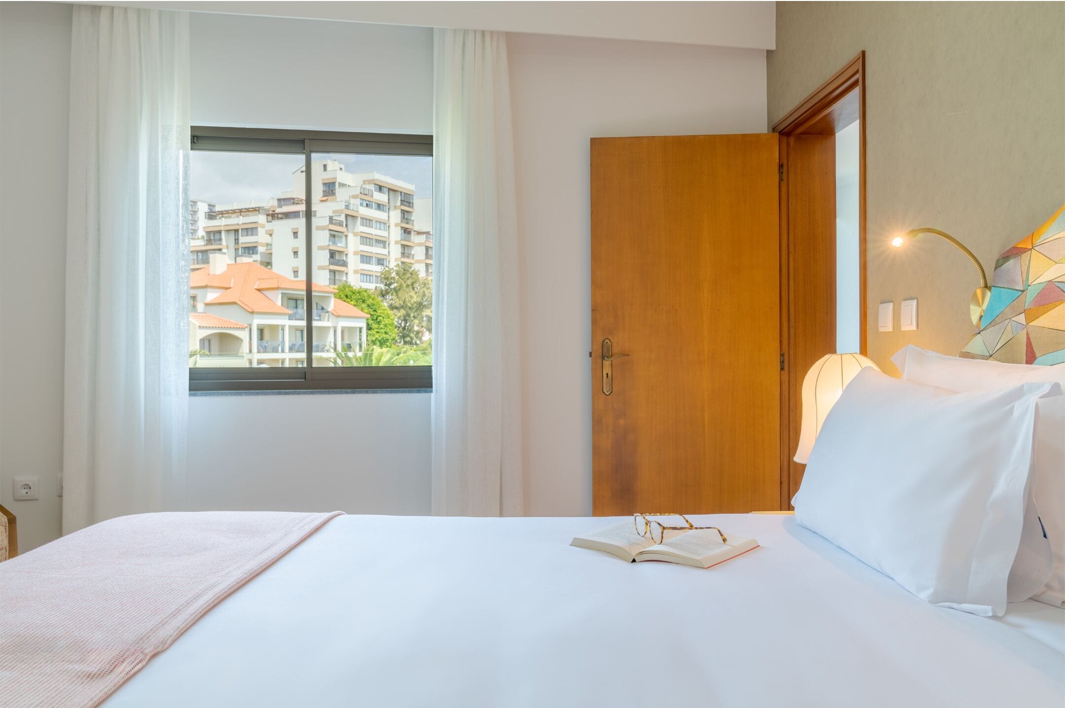 Une chambre d’hôtel avec un lit, une fenêtre avec des rideaux, une porte en bois et une lampe fixée au mur.