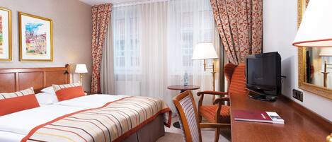 Habitación de hotel con cama, escritorio con televisor, silla y lámpara. Hay cortinas y una ventana con vistas.