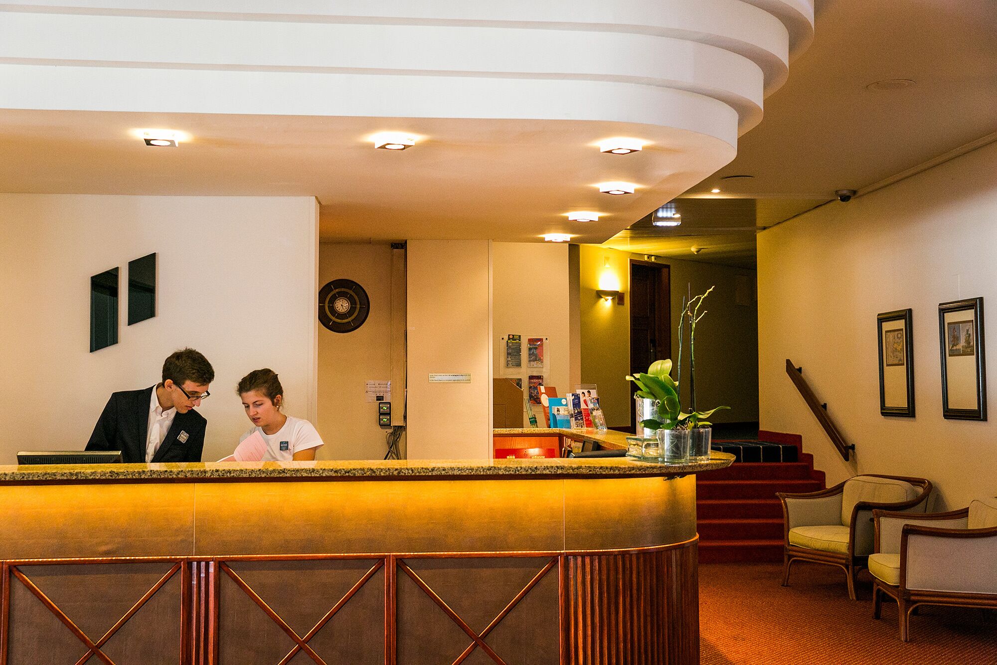 Zona de recepción de hotel con un mostrador curvo, un reloj, cuadros enmarcados en la pared y una escalera que sube a otro piso.