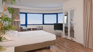 Een moderne hotelkamer met een groot bed, een televisie, een bureau en uitzicht op de stad door grote ramen.