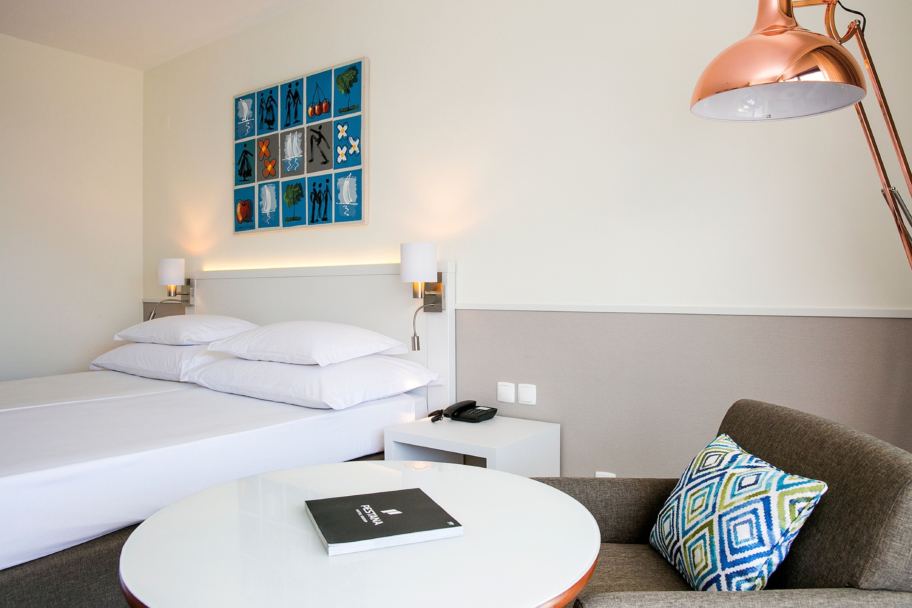 Habitación de hotel con cama, mesita de noche blanca con lámpara, una pequeña mesa con un libro, sofá con cojín estampado y una pared con una obra de arte azul.