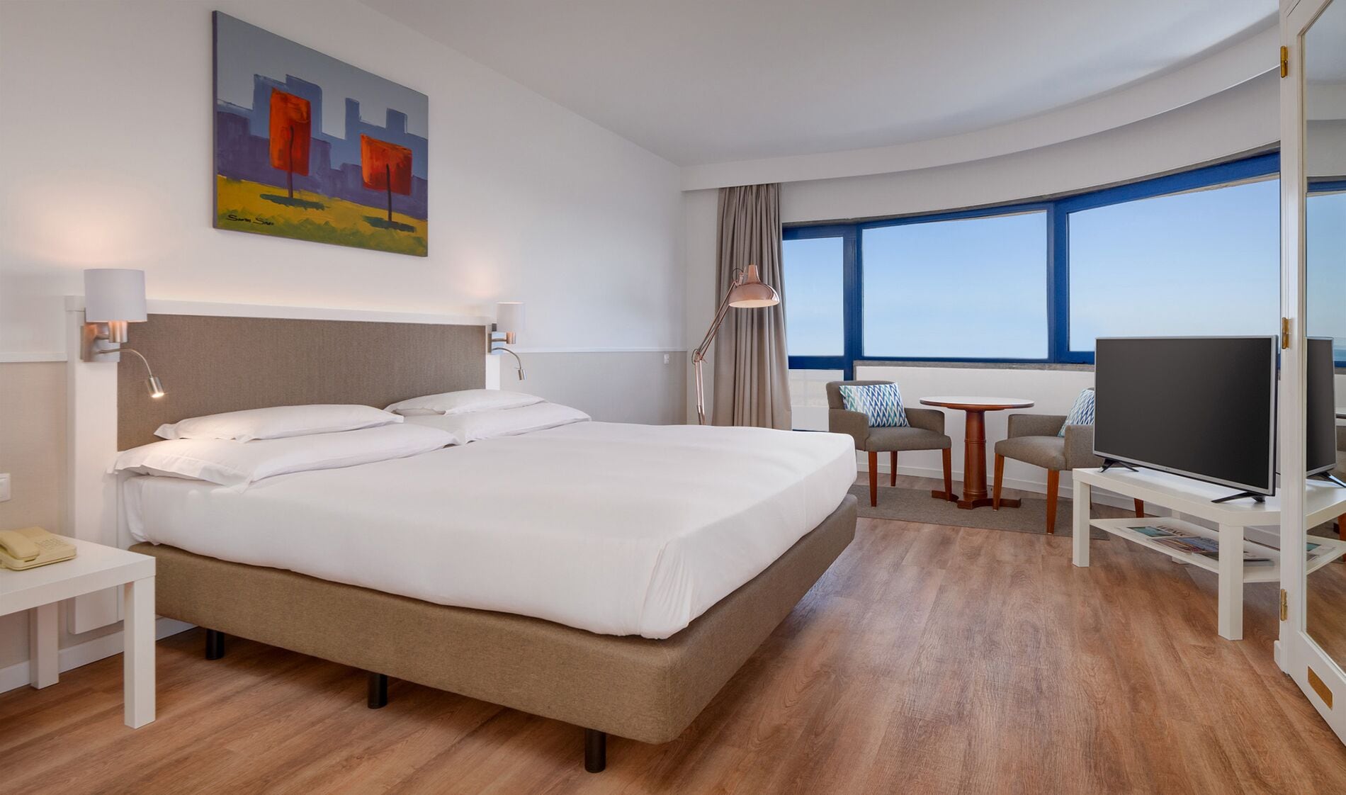Habitación de hotel con una cama grande, un escritorio con televisor, una silla y un cuadro en la pared.