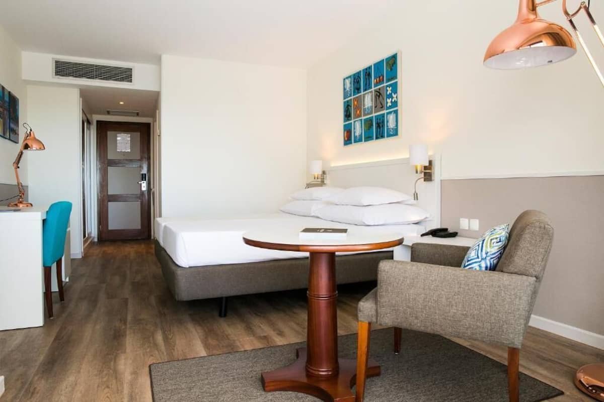 Une chambre d’hôtel comprenant un lit, une table ronde en bois, une chaise, un bureau avec une chaise, une lampe et une œuvre d’art murale.