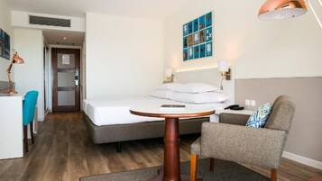 Quarto de hotel com uma cama, uma mesa redonda de madeira, uma cadeira, uma escrivaninha com cadeira, um abajur e uma obra de arte na parede.