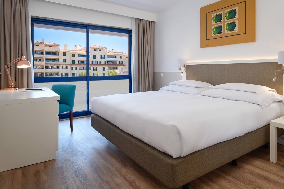 Une chambre d’hôtel avec un grand lit, un bureau avec une lampe, une chaise et une vue sur les immeubles par la fenêtre.