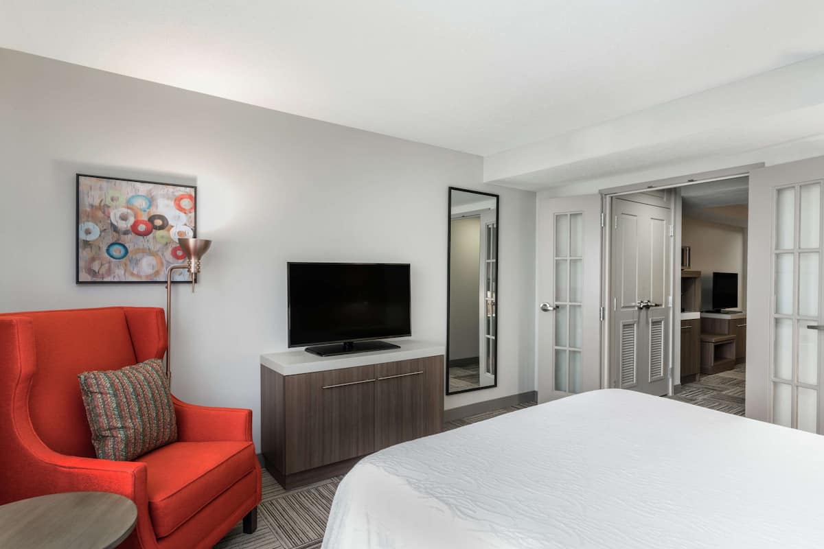 A modern hotel room with a large bed, a red armchair, a flat-screen TV, a mirror, and a wardrobe.