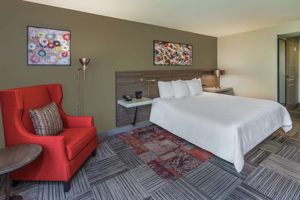 A hotel room with a large bed, a red armchair, a small table, two paintings on the walls, and a floor lamp.