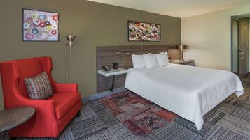 Habitación de hotel con una cama grande, un sillón rojo, una mesita, dos cuadros en las paredes y una lámpara de pie.