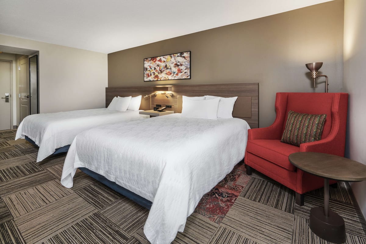 A hotel room with two beds, a red armchair, a small table, and a wall-mounted lamp.