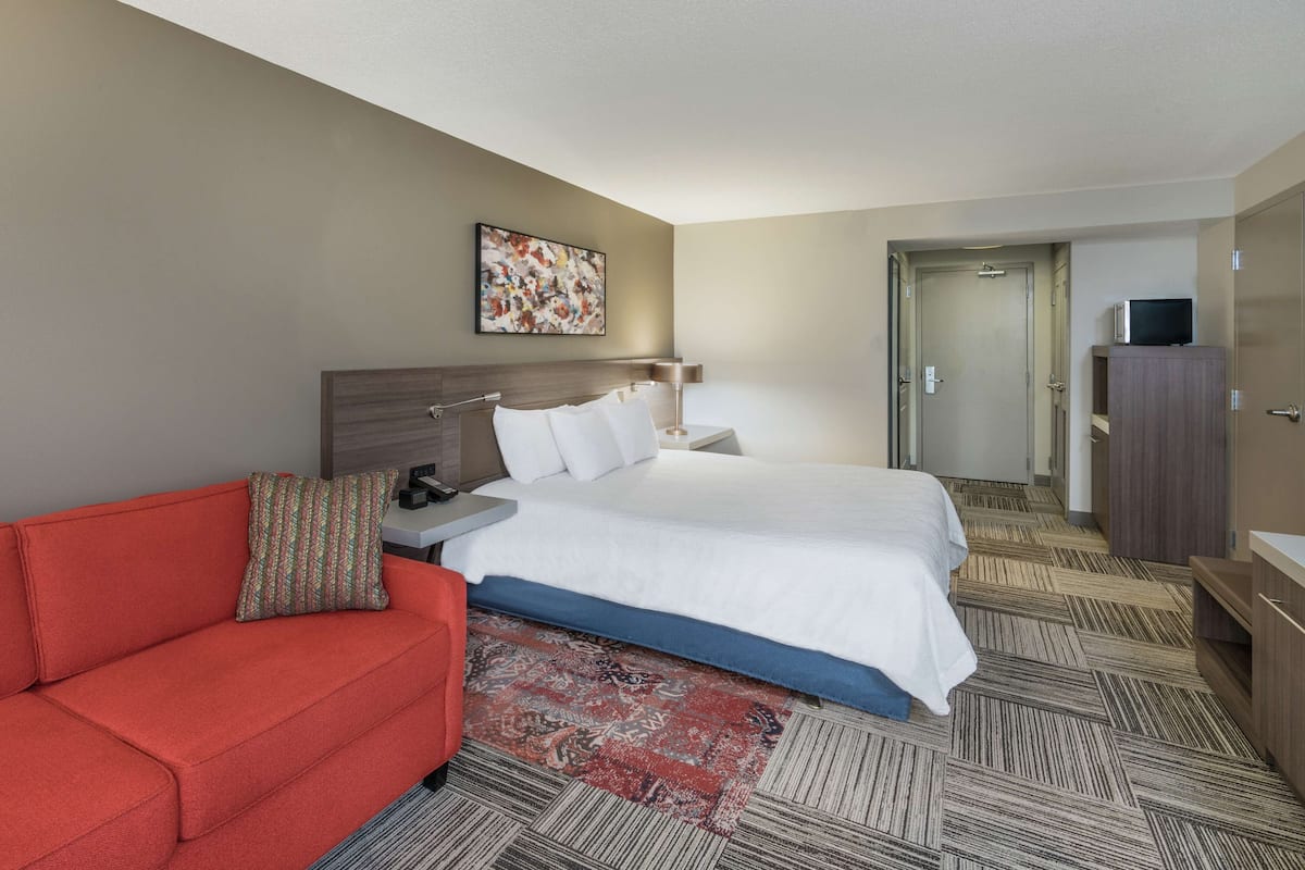 A hotel room with a bed, a red sofa, a nightstand, a lamp, a television on a cabinet, and a patterned rug.