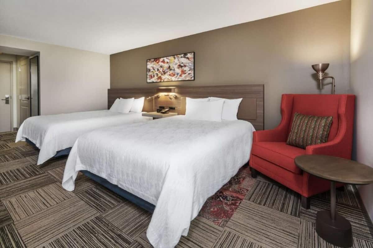 A hotel room with two beds, a red armchair, a small table, and a wall-mounted lamp.