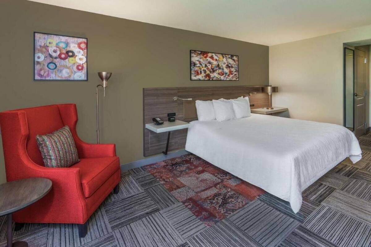 A hotel room with a bed, a red armchair, a small table, a floor lamp, and two abstract paintings on the walls.