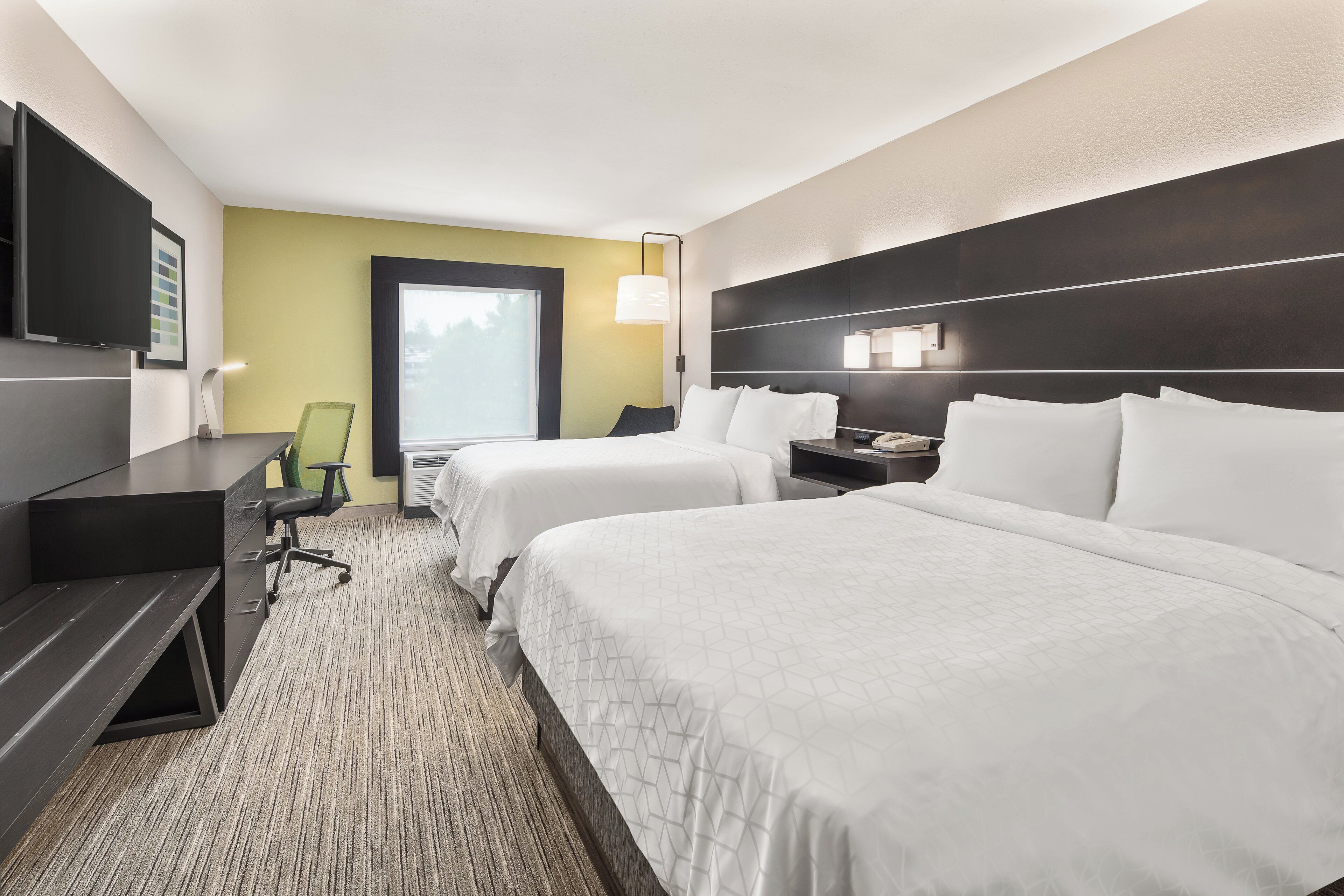 A hotel room with two beds, a desk, a chair, a TV, and a window.