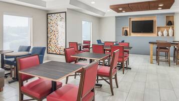 A hotel dining area with multiple tables and chairs, a large window, and a flat-screen TV mounted on the wall.