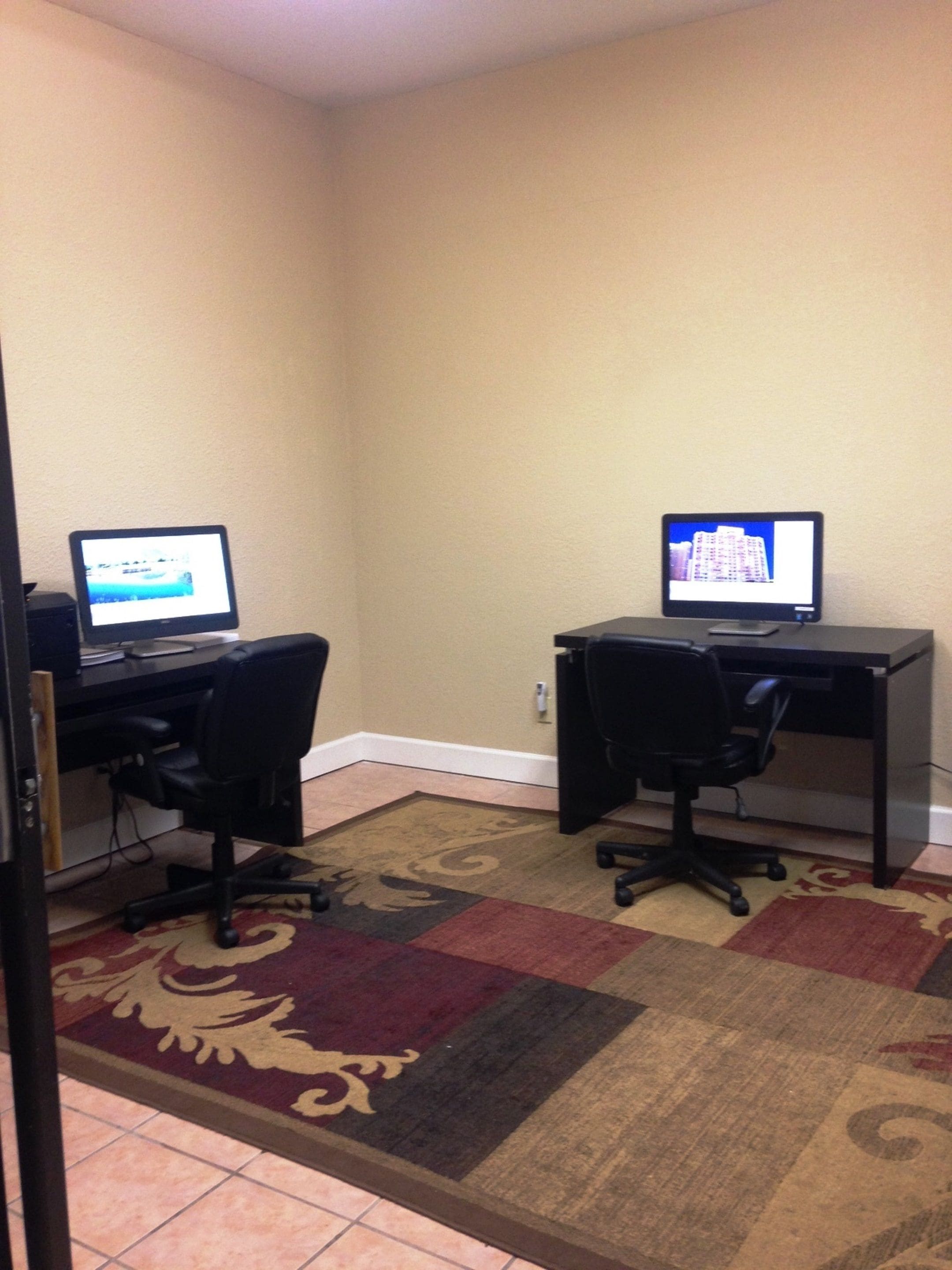 Una habitación con dos escritorios, cada uno con una computadora y una silla. El piso tiene una alfombra estampada.