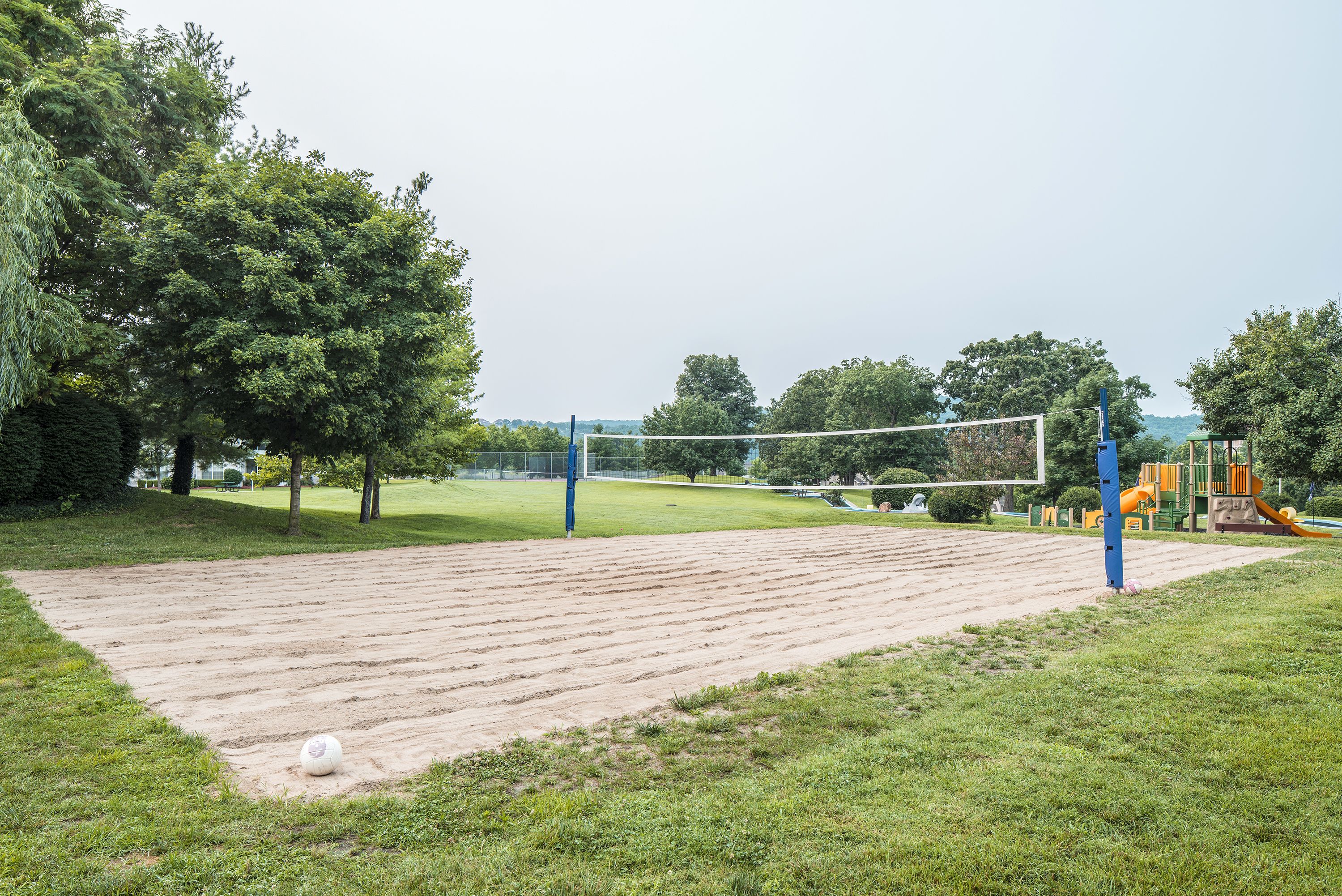 A volleyball court with a net, surrounded by trees and a playground.