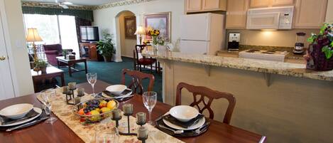 Un comedor con una mesa lista para cuatro personas, un televisor al fondo y una zona de cocina con microondas y estufa.