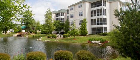 A multi-story building with a pond in front, surrounded by greenery and a small bridge.