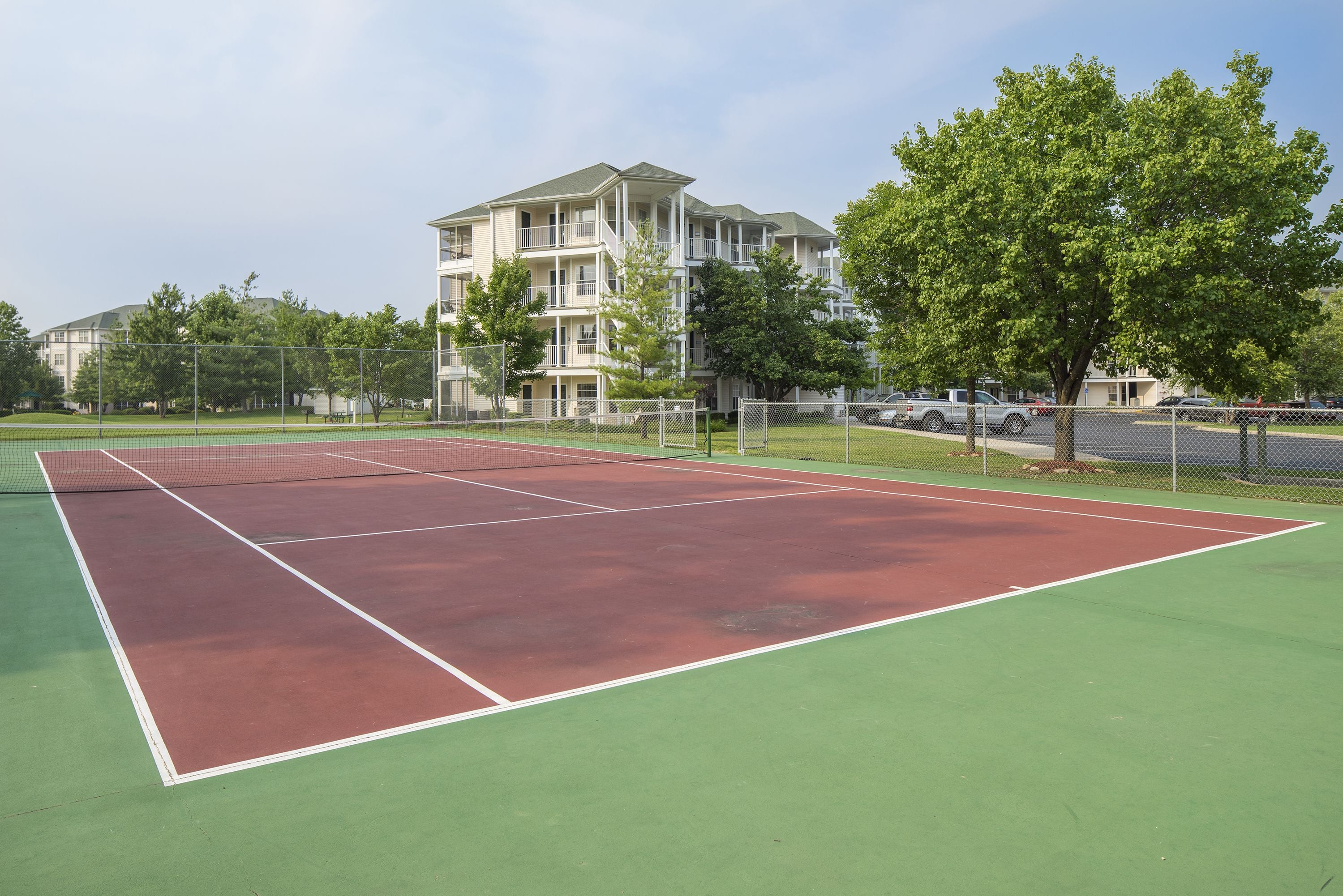 A tennis court with a net, surrounded by a fence, and a building with multiple balconies in the background.