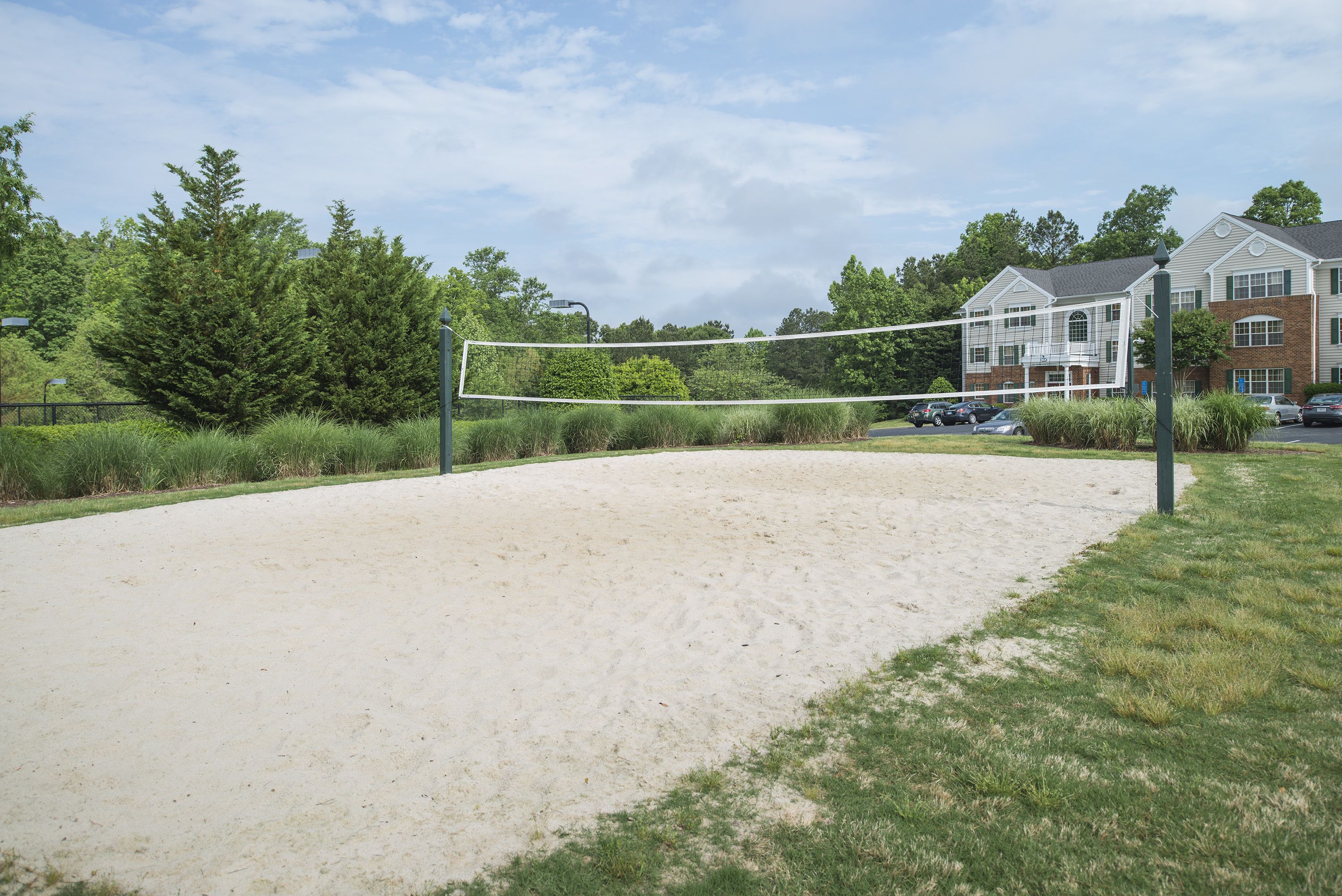 Un terrain de volleyball de sable avec un filet, entouré d’herbe et d’arbres, avec un bâtiment en arrière-plan.