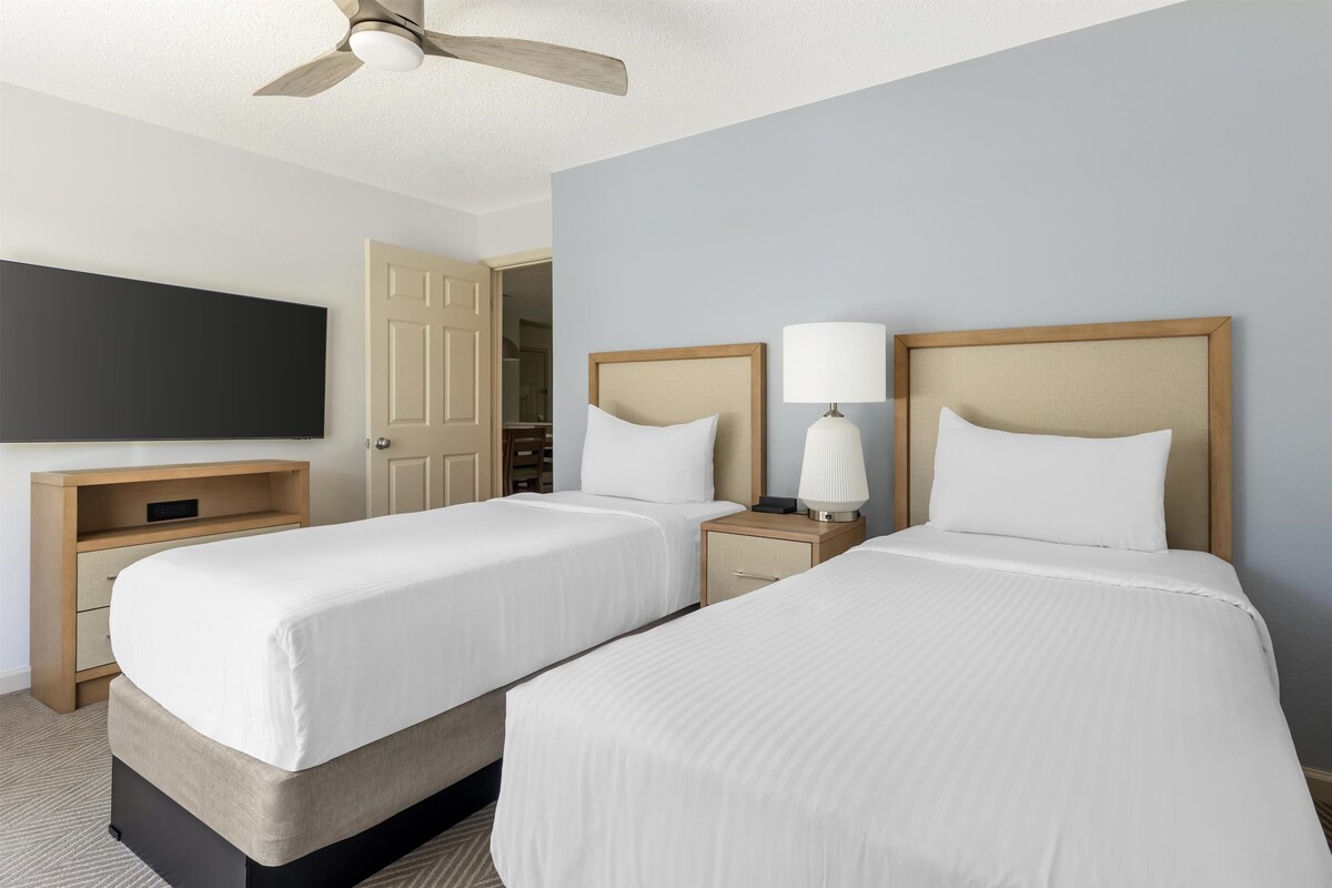 Habitación de hotel con dos camas, televisión, ventilador de techo y puerta que da a un baño.