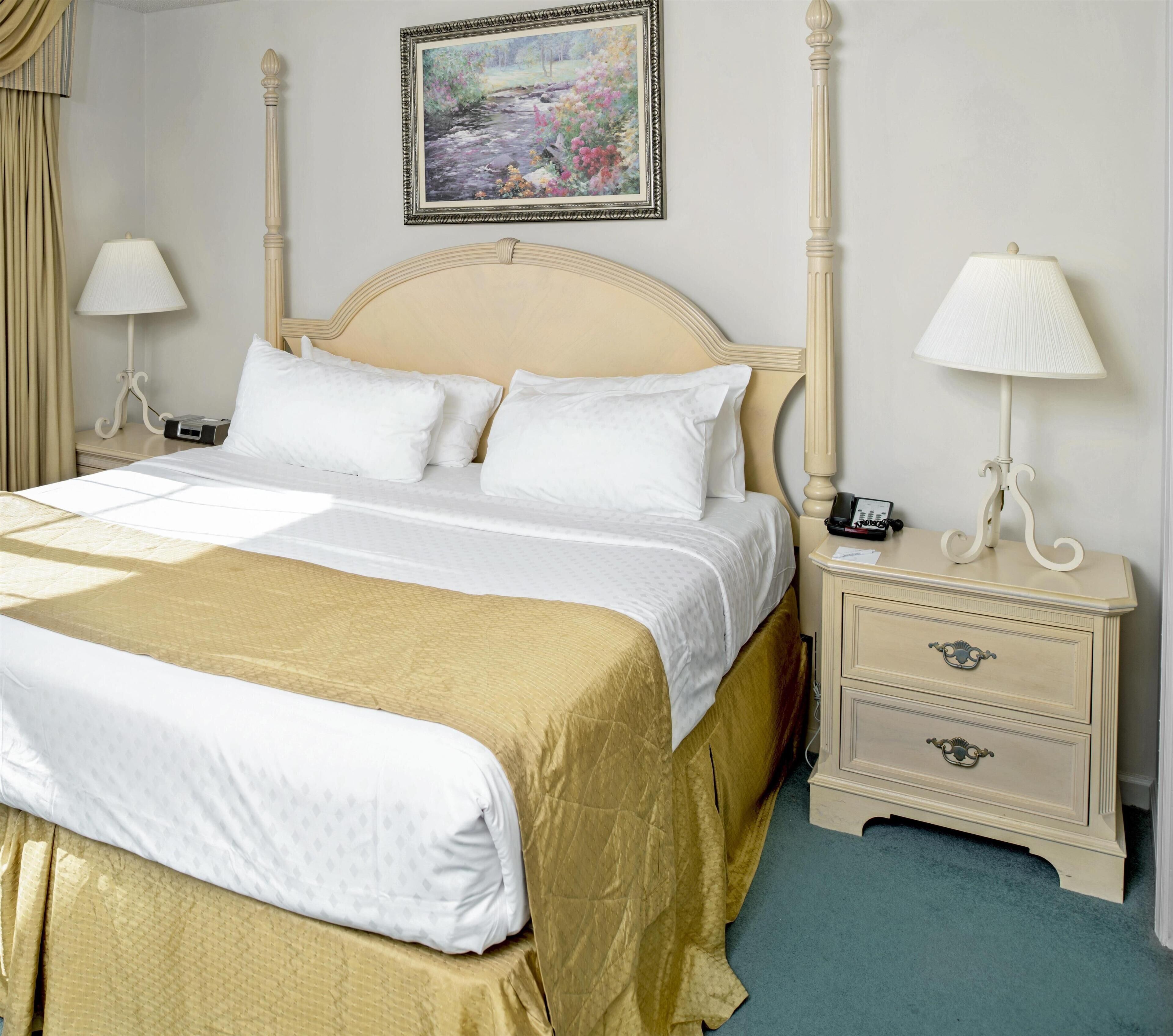A neatly made bed with a headboard, two bedside tables with lamps, a painting on the wall, and a blue carpet.