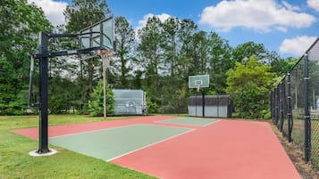 An outdoor basketball court with two hoops, surrounded by trees and a fenced area.