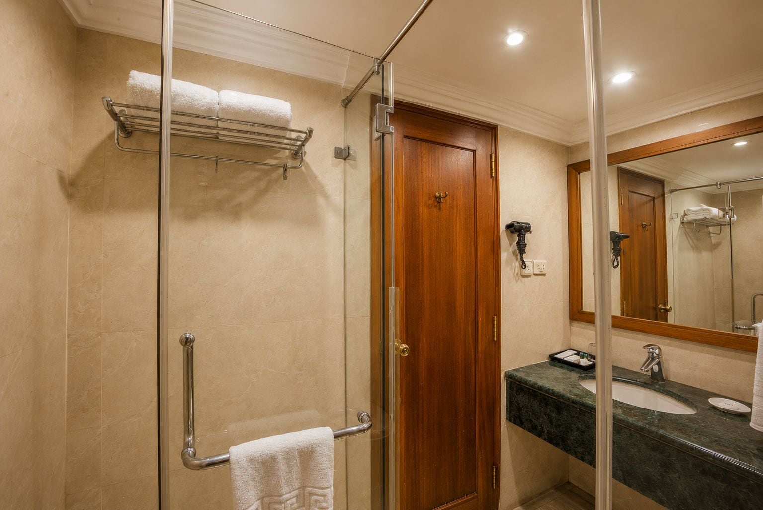 A hotel bathroom with a glass-enclosed shower, a wooden door, a sink with a marble countertop, and a towel rack.
