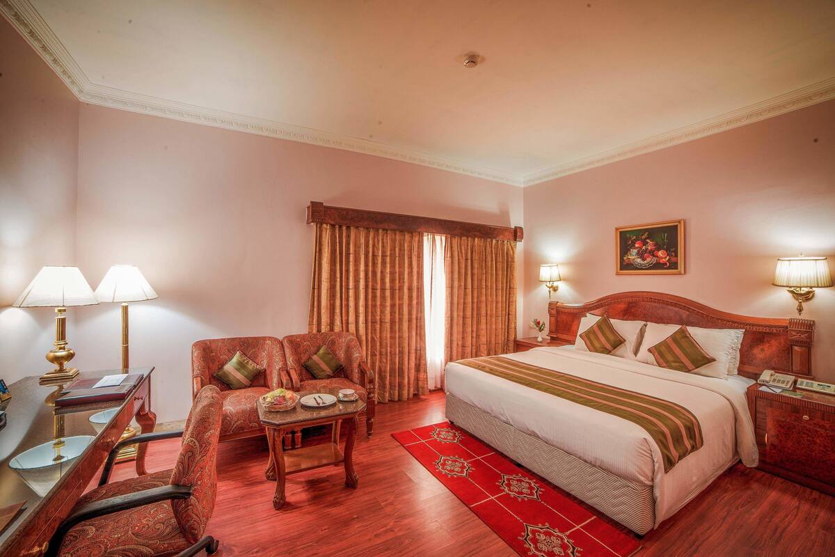 Quarto de hotel com cama, escrivaninha com luminária, cadeira, mesa com um vaso e um tapete vermelho.