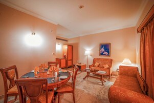 A hotel room with a dining table, chairs, a sofa, a lamp, and a painting on the wall.