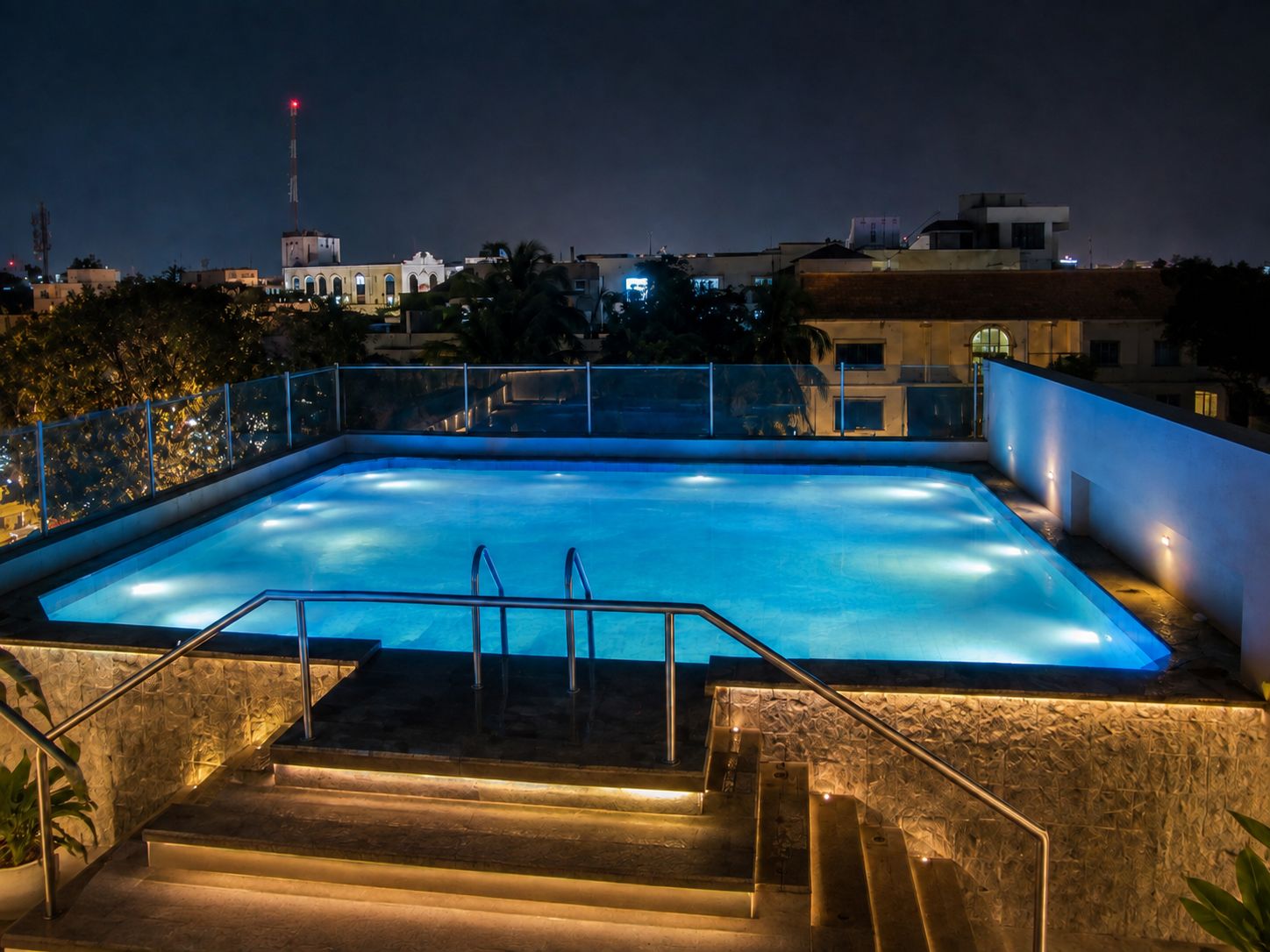A rectangular swimming pool with a glass barrier and steps leading down to it, surrounded by buildings.