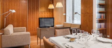 A modern hotel room with a wooden interior, a flat-screen TV, a dining area set for four, and a bookshelf filled with books.