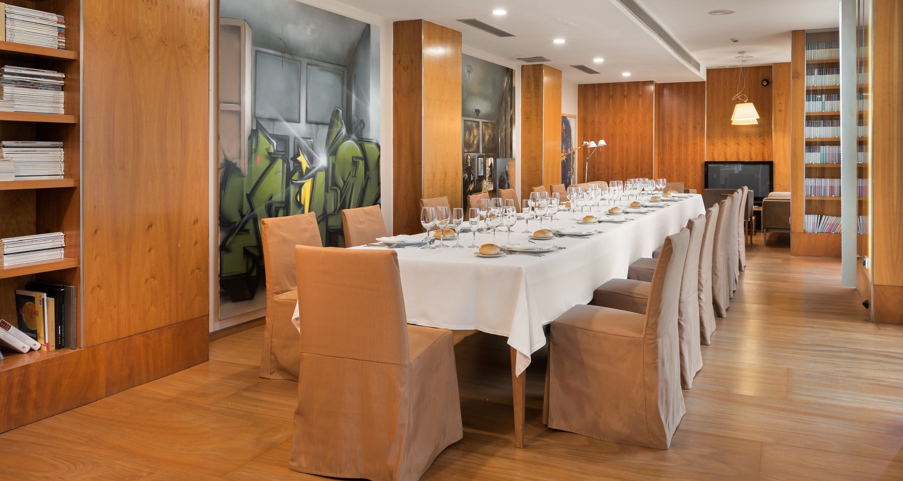 Una larga mesa de comedor dispuesta con sillas, rodeada de estanterías de madera y un mural.