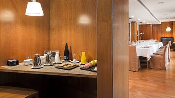 Disposición de un bufé de desayuno con diversos alimentos y bebidas, una larga mesa de comedor con sillas y un televisor al fondo.