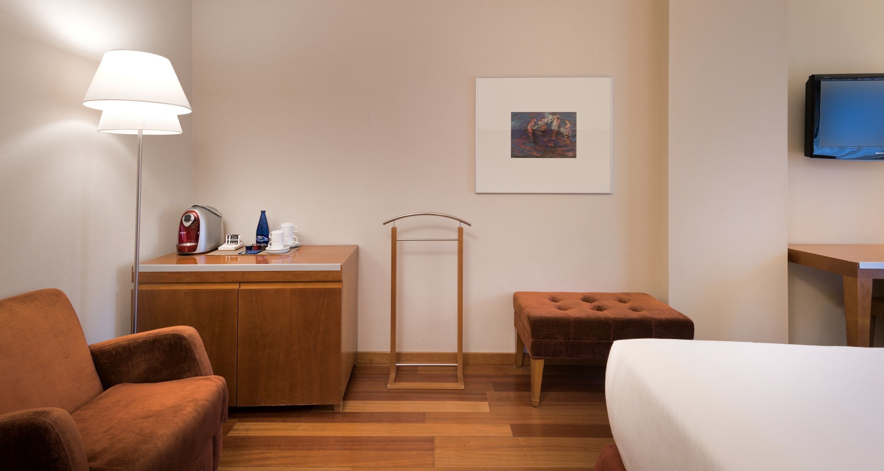Habitación de hotel con cama, escritorio con lámpara, silla, una mesita y un cuadro en la pared.
