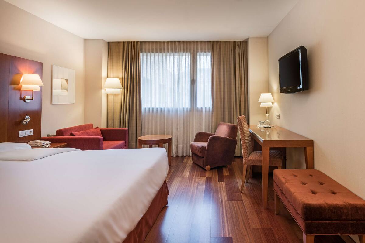 A hotel room with a bed, two armchairs, a small table, a desk, a television, and a window with curtains.