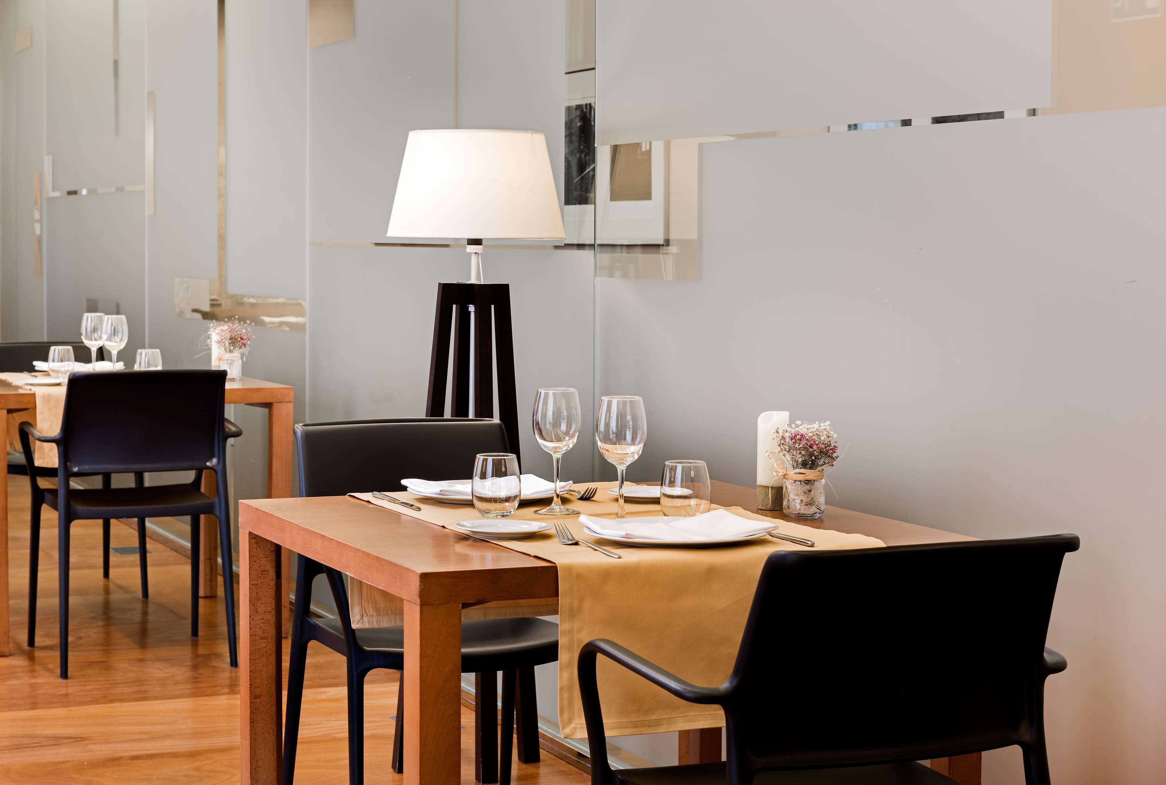 Un comedor con una mesa de madera dispuesta para dos comensales, sillas negras, una lámpara de mesa y un florero con flores.