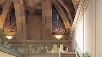 A grand staircase with a glass railing, leading to an upper level with ornate ceiling decorations and a view of the cityscape.