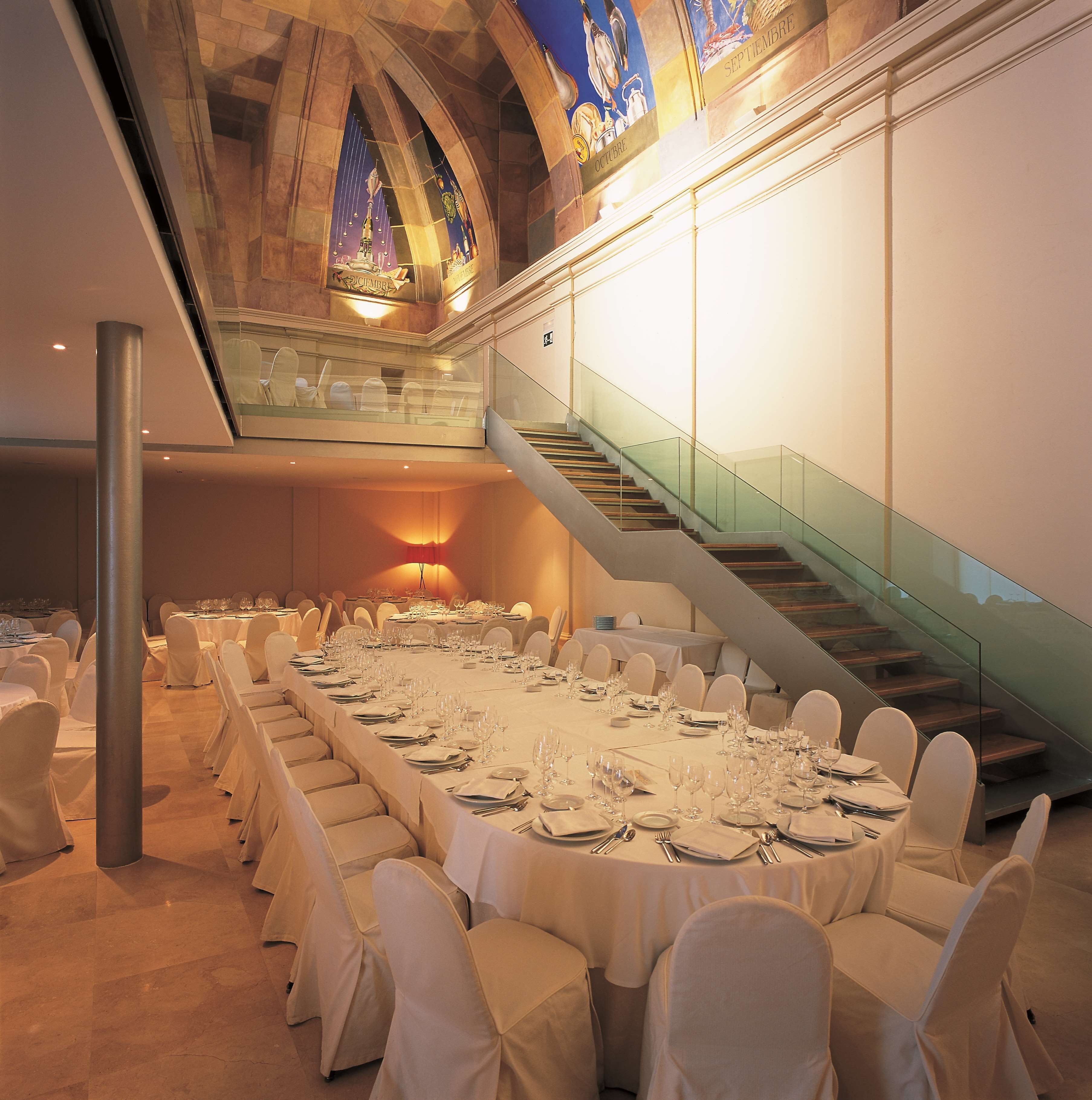A banquet hall with a long table set for a formal event, a staircase with glass railings, and a stained glass ceiling.