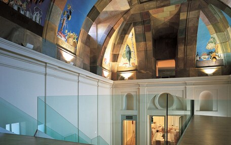 A grand interior with a high arched ceiling, a staircase leading to a dining area, and a mural depicting agricultural scenes.